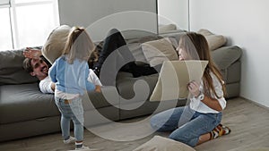 Little cute girl having funny pillow fight with dad on couch in living room and mom is sitting on the floor playing with