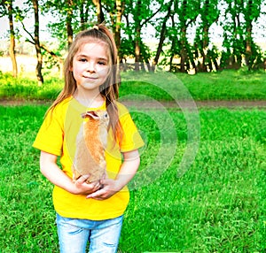 A little cute girl holds a real red rabbit in her arms against the background of green plants. Place for inscription