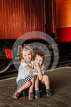 Little cute girl hugs a boy, they are sitting on a black suitcase on a peron at the station and waiting for the train. A long