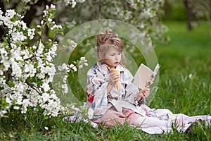 Little cute girl with ice cream reading a book  in the park. Child outdoors in blossoming spring garden