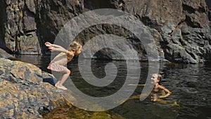 Little cute girl jumping in water of mountain lake to her mother