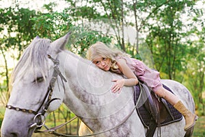 Little cute girl with light curly hair in a straw hat riding a horse at sunset