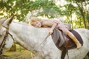 Little cute girl with light curly hair in a straw hat riding a horse at sunset