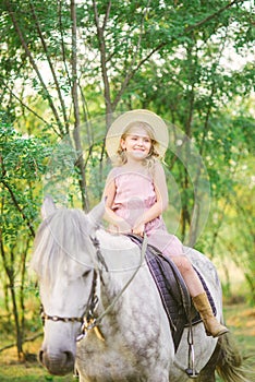 Little cute girl with light curly hair in a straw hat riding a horse at sunset