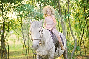 Little cute girl with light curly hair in a straw hat riding a horse at sunset