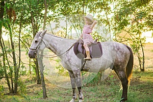 Little cute girl with light curly hair in a straw hat riding a horse at sunset