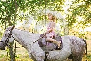 Little cute girl with light curly hair in a straw hat riding a horse at sunset