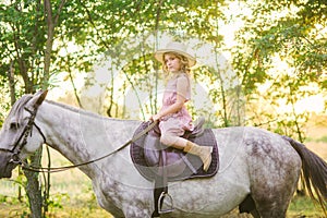 Little cute girl with light curly hair in a straw hat riding a horse at sunset