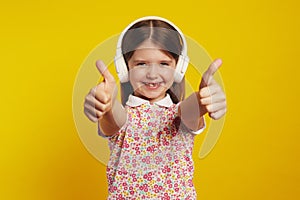 Little cute girl listening music in white headphones and showing thumbs up
