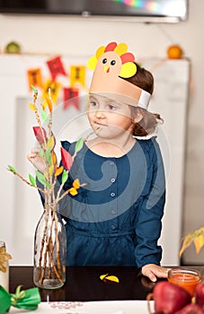 Little cute girl in paper turkey hat glues paper leaves on branches for celebrating Thanksgiving day. Diy craft art