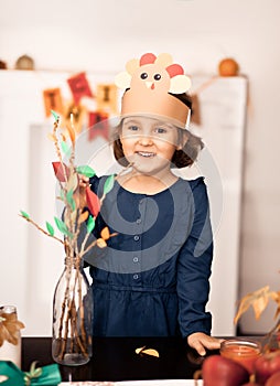 Little cute girl in paper turkey hat glues paper leaves on branches for celebrating Thanksgiving day. Diy craft art
