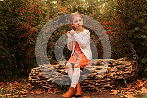 Little cute girl with pigtails sits on a textured stone among autumn foliage