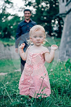 Little cute girl in pink dress standing in front of her father on green grass near old stone walls