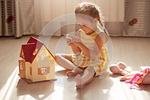 Little cute girl playing with her small kitty while sitting on floor in nursery. Children play with wooden toy house. Toddler kid