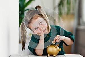 Little cute girl putting money into gold piggy bank at table indoors. Primary financial education