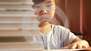 A little cute girl reading a book sits on table at home. Stay at home to quarantine from coronavirus pandemic. Homeschool concept.