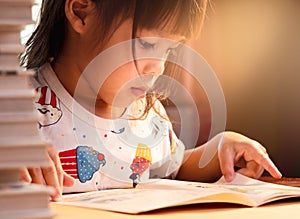 A little cute girl reading a book sits on table at home. Stay at home to quarantine from coronavirus pandemic. Homeschool concept.