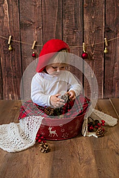 A little cute girl in a red plaid skirt and a red felt beret plays with cones and Christmas toys in a room decorated for Christmas