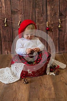 A little cute girl in a red plaid skirt and a red felt beret plays with cones and Christmas toys in a room decorated for Christmas