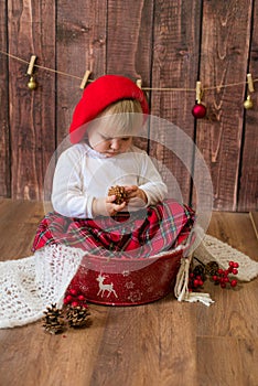 A little cute girl in a red plaid skirt and a red felt beret plays with cones and Christmas toys in a room decorated for Christmas