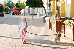 A little cute girl in red and white striped pants is walking with her French bulldog dog