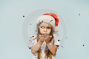 Little cute girl in santa hat blows confetti into the camera. Christmas and New Year