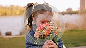 Little cute girl sits on a park bench near small lake with a bouquet of flowers.