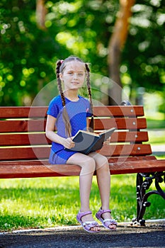 Little cute girl is sitting on a bench and is reading a book