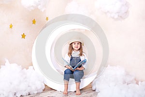 Little cute girl sitting on the moon with clouds and stars with a book in her hands and reading. The girl is learning to read. Rea