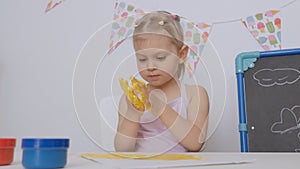 A little cute girl sitting at the table in the children`s room drawing with finger paints stained her hands in yellow