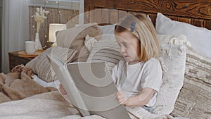 Little cute girl small daughter kid child baby toddler preschooler sits in bed in cozy bedroom interesting reads book