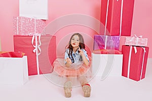 Little cute girl in tulle skirt expressing to camera on white floor suround colorful giftboxes on pink background. Sweet