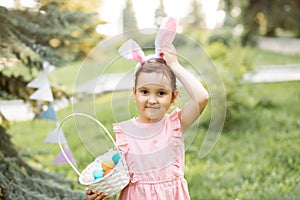 Little cute girl wear bunny ears holding basket with colorful painted eggs on Easter egg hunt in park