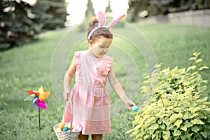 Little cute girl wear bunny ears holding basket with colorful painted eggs on Easter egg hunt in park