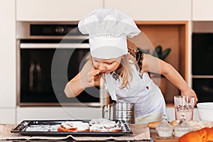 A little cute girl with a white chef hat tastes pastries in the kitchen