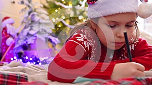 Little cute girl writing a letter to Santa Claus. A child in a hat and a red sweater at home near the Christmas tree.