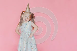 A little cute happy girl in birthday cap  grimaces with happy face on a pink background.