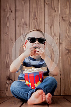 Little cute kid baby boy 2-3 years old , 3d cinema glasses holding bucket for popcorn, eating fast food on wooden background. Kids