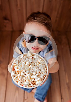 Little cute kid baby boy 2-3 years old , 3d imax cinema glasses holding bucket for popcorn, eating fast food on wooden background