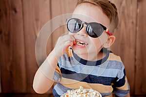 Little cute kid baby boy 2-3 years old , 3d imax cinema glasses holding bucket for popcorn, eating fast food on wooden background