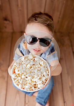 Little cute kid baby boy 2-3 years old , 3d imax cinema glasses holding bucket for popcorn, eating fast food on wooden background