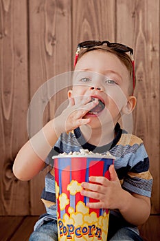 Little cute kid baby boy 2-3 years old , 3d imax cinema glasses holding bucket for popcorn, eating fast food on wooden background