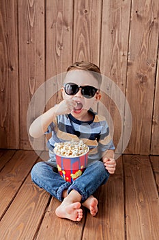 Little cute kid baby boy 2-3 years old , 3d imax cinema glasses holding bucket for popcorn, eating fast food on wooden background