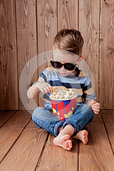 Little cute kid baby boy 2-3 years old , 3d imax cinema glasses holding bucket for popcorn, eating fast food on wooden background