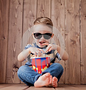 Little cute kid baby boy 2-3 years old , 3d imax cinema glasses holding bucket for popcorn, eating fast food on wooden background