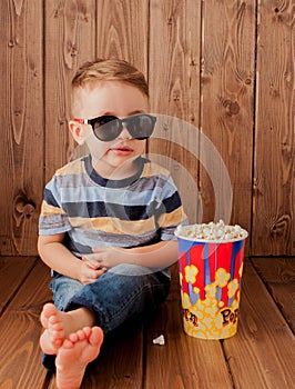 Little cute kid baby boy 2-3 years old , 3d imax cinema glasses holding bucket for popcorn, eating fast food on wooden background