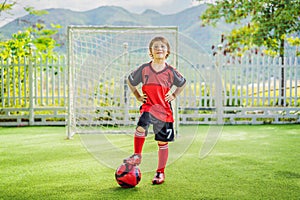 Little cute kid boy in red football uniform playing soccer, football on field, outdoors. Active child making sports with