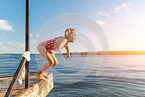 Little cute kid girl in swimsuit have fun enjoy pretend flying jumping from pier dock in clean blue water sea river or