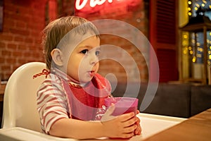 A little cute kid sits in a highchair, holds a glass of juice with his hands and drinks from a straw. in the background