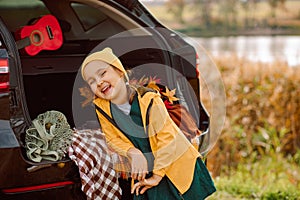 Little cute laughing girl standing near by on open car trunk. Kid resting with her family in the nature. Autumn season